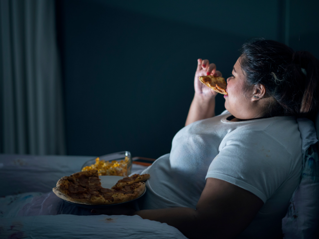 Person eating pizza at night time while watching TV. Learning about why do we overeat at night?