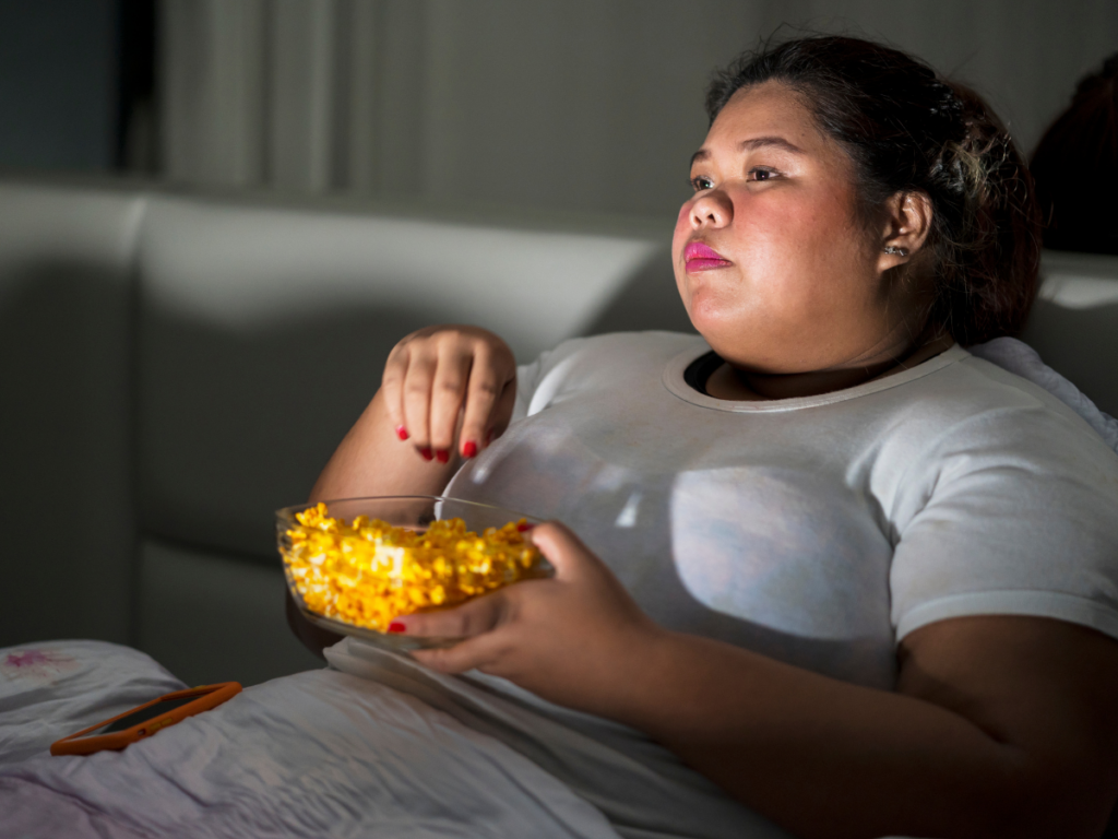 Woman sitting on the couch watching TV and snacking learning about signs of comfort and emotional eating