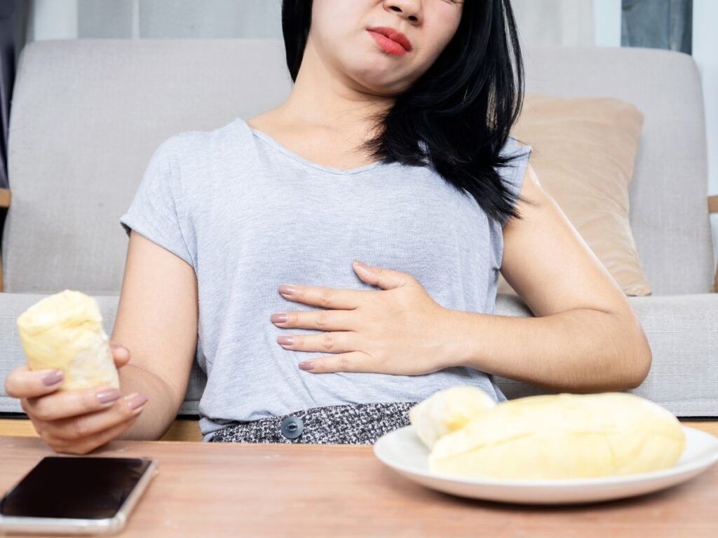 Woman with hand on stomach with plate of food in front of her feeling uncomfortable from overeating because of the clean plate club