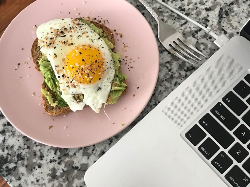 Avocado toast with egg on it on a pink plate beside a laptop showing you how to eat intuitively when you're busy