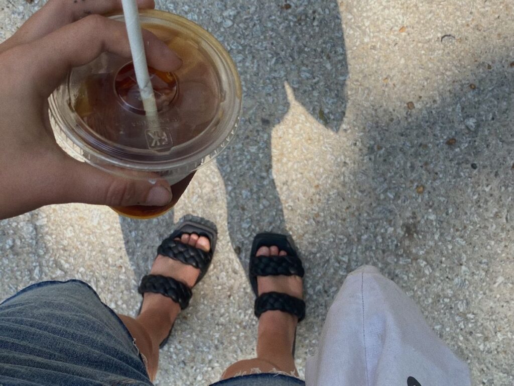 Person holding iced coffee looking down at their feet wearing black sandals and is trying to practice intuitive eating for a busy life